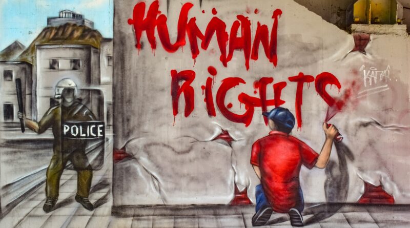 Un home grafitejant "drets humans" en una paret mentre un policia el mira de forma amenaçant.